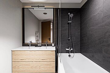 Stylish bathroom featuring a light wood double vanity, large mirror, and a sleek dark tiled shower/tub combo with glass partition.