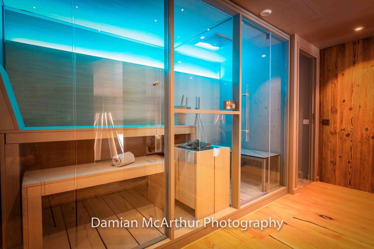 Modern glass-walled sauna and steam room illuminated by vibrant blue lighting, featuring wooden benches and floors.