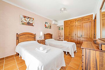 A bright bedroom with two single beds, white bedding, wooden furniture including a wardrobe and dresser, and light pink walls with paintings.