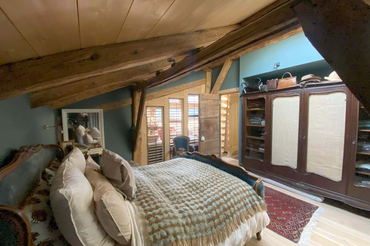 A cozy attic bedroom with rustic wooden beams, slanted ceiling, and blue walls. Features a large bed, vintage wardrobe, and shuttered windows.