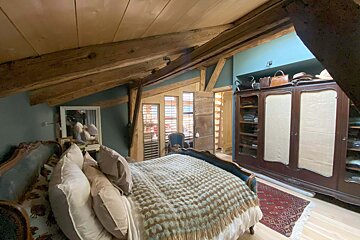 A cozy attic bedroom with rustic wooden beams, slanted ceiling, and blue walls. Features a large bed, vintage wardrobe, and shuttered windows.