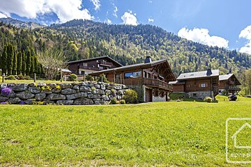 Wooden chalets on a green lawn with a stone wall in the foreground, backed by a lush, tree-covered mountain under a blue sky.