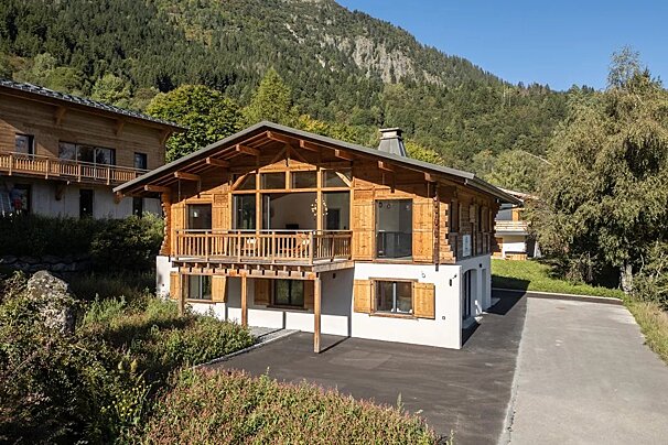A modern two-story wooden chalet with a balcony, set against a backdrop of forested mountains.