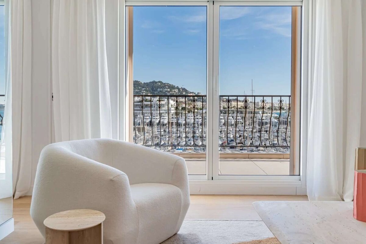 A modern room with a white armchair and sheer curtains, offering a scenic view from a balcony over a marina filled with yachts and distant hills.