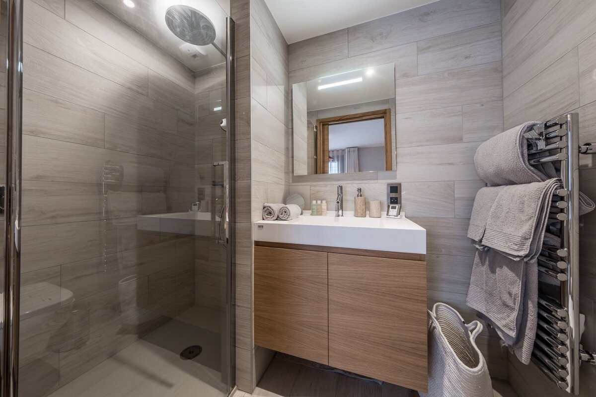 Modern bathroom with a glass shower, wooden vanity, tiled walls, and a chrome towel warmer with grey towels.
