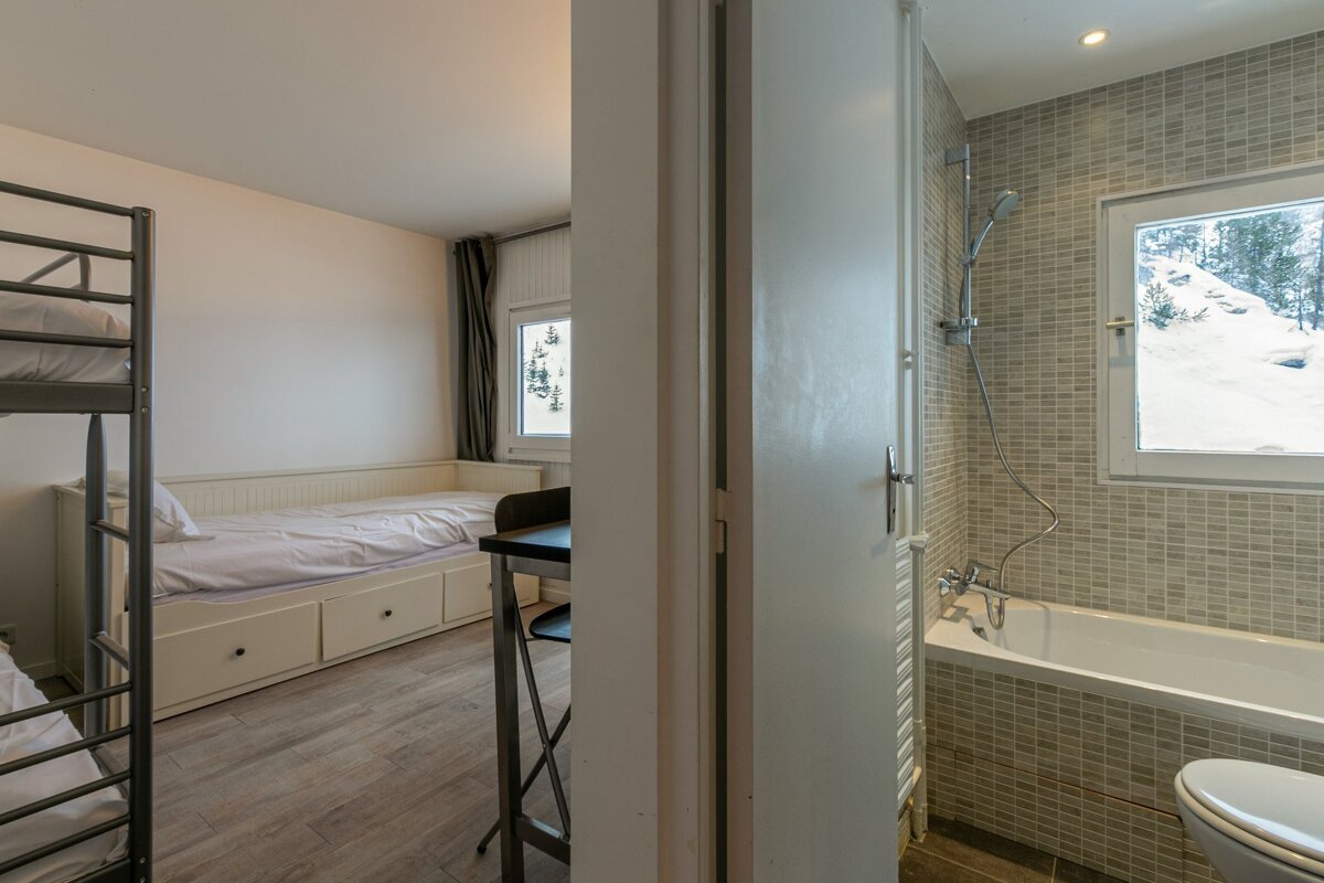 A bedroom with bunk beds and a daybed, next to a tiled bathroom with a tub/shower. Both rooms have windows looking out to a snowy mountain view.