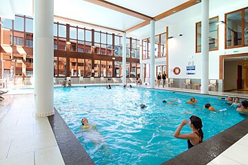 People enjoy a bright, spacious indoor swimming pool with large windows revealing buildings outside.