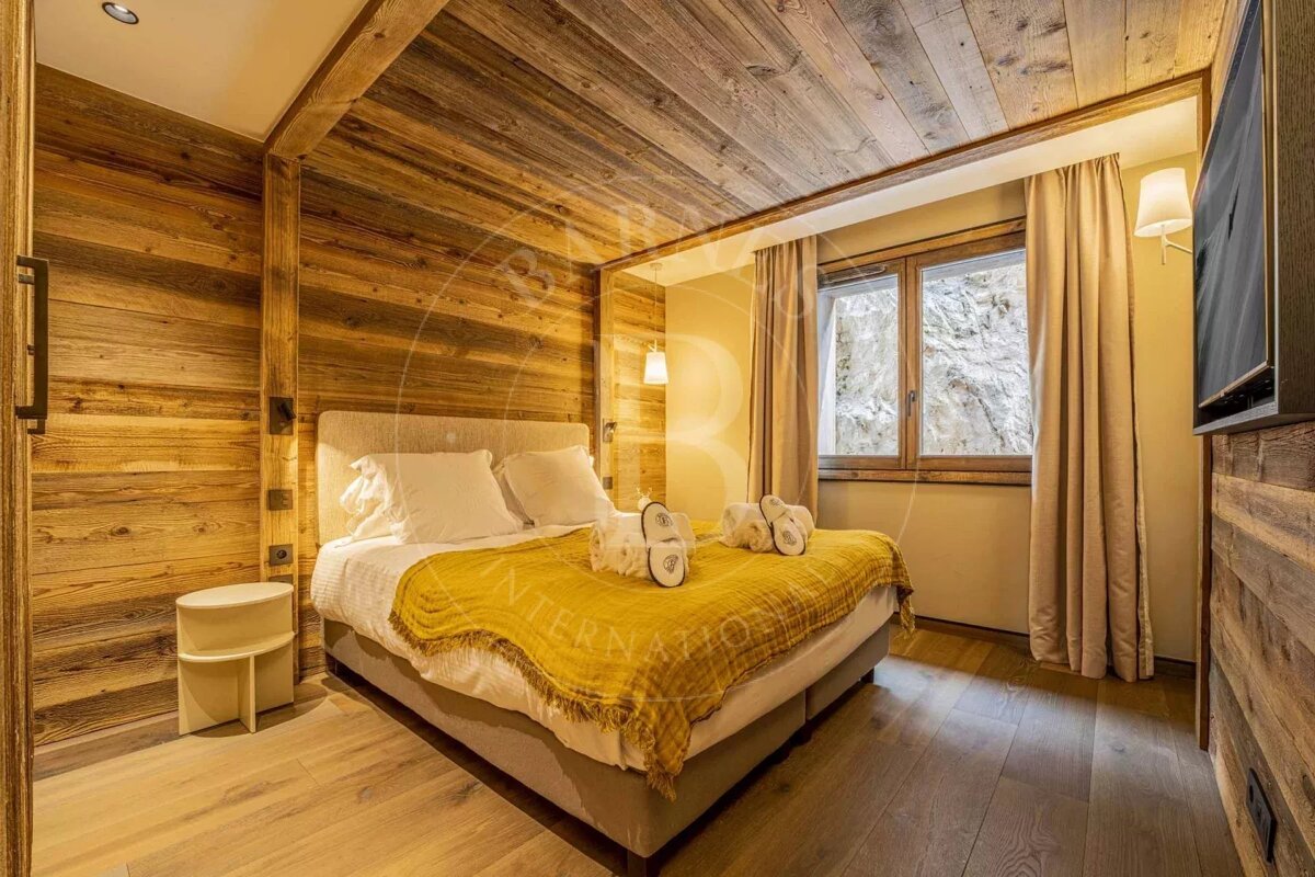 A cozy, rustic bedroom featuring wood-paneled walls and ceiling, a bed with a yellow throw, and a window overlooking a rocky outdoor view.
