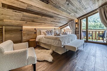 Cozy alpine chalet bedroom with rustic wood paneling, a white bed, a plush armchair, and glass doors leading to a scenic balcony.