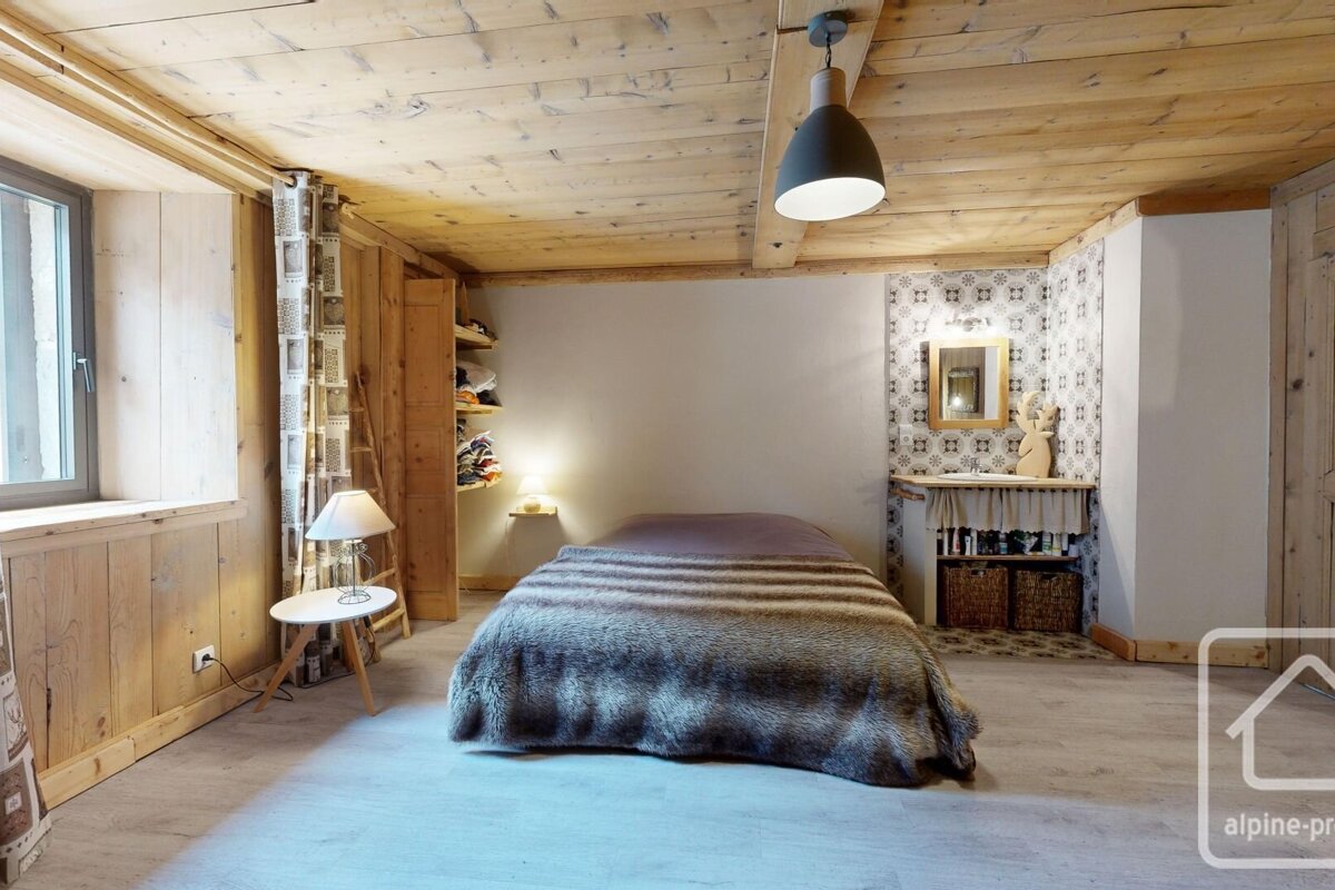 A rustic bedroom featuring a wooden ceiling, exposed wood paneling, a bed with a furry blanket, and a compact corner vanity with patterned tiles and a mirror.
