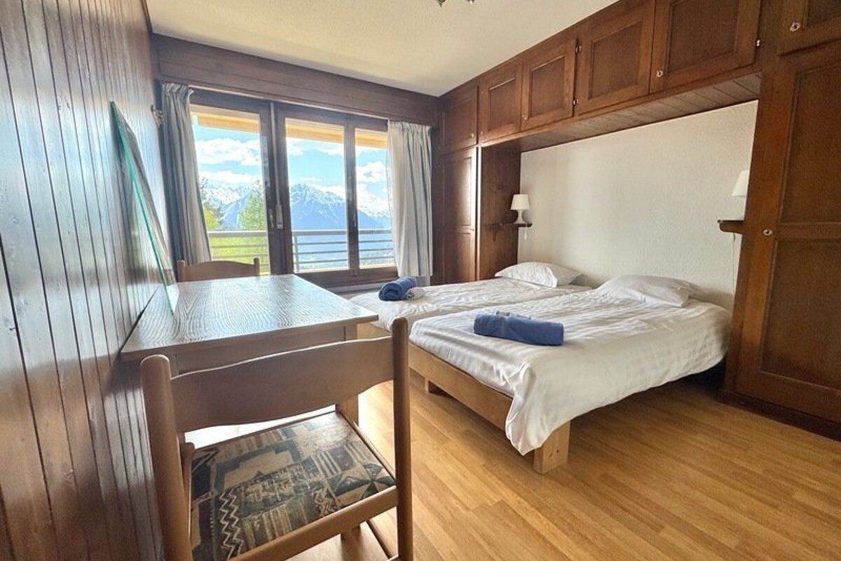 Cozy wood-paneled bedroom featuring twin beds, a dining table, and a balcony offering a scenic mountain view.