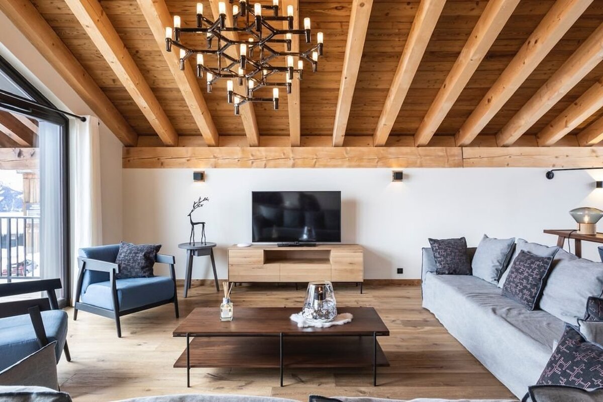 Modern chalet living room featuring exposed wooden ceiling beams, a large statement chandelier, comfortable seating, and a TV console.