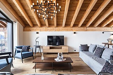 Modern chalet living room featuring exposed wooden ceiling beams, a large statement chandelier, comfortable seating, and a TV console.