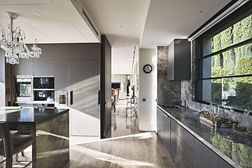 A chic, modern kitchen boasts dark wood cabinetry, marble counters, a chandelier, and a large window overlooking lush greenery, with sunlight streaming inside.