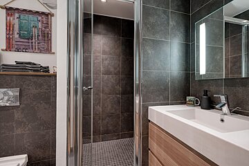 Modern bathroom with dark grey tiles, glass shower, white sink with wooden vanity, and a mirror. Ski-themed art hangs on the wall.