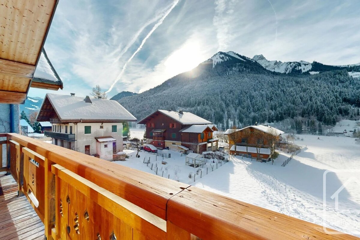 View from a wooden balcony overlooking snow-covered alpine chalets and forested mountains under a bright winter sky.