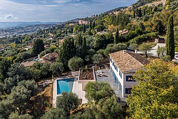 Aerial view of a white house with a pool, nestled in a lush, green, olive tree-filled hillside overlooking a distant town.