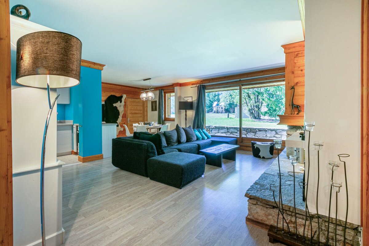 A modern rustic living room with a dark sectional sofa, bright blue wall, and wood accents. A large window offers views of a green outdoor space.