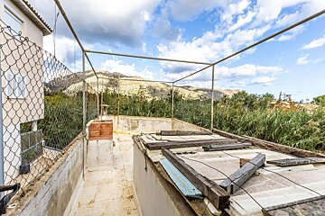 A weathered rooftop terrace with a metal frame, overlooking overgrown reeds and distant mountains under a cloudy sky.