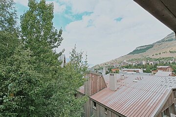 A scenic view of a mountain town from a window, with lush green trees, rustic wooden buildings, and a dry mountain under a partly cloudy sky.