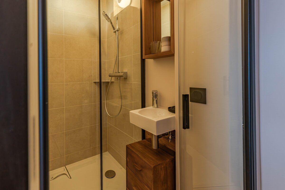 A compact modern bathroom featuring a glass-enclosed shower with beige tiles, a small sink, and wooden storage.