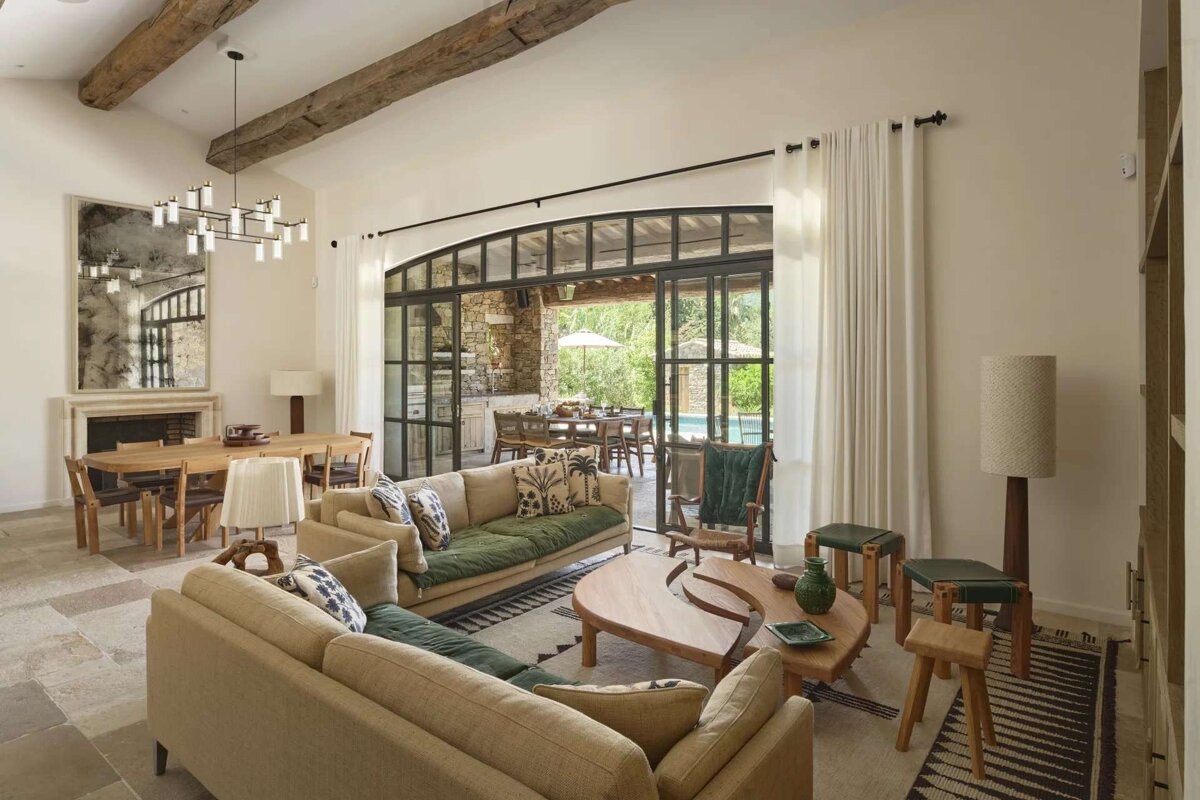 Bright, open living/dining area with rustic beams, modern chandelier, and large glass doors to an outdoor patio and pool. Earth tones.