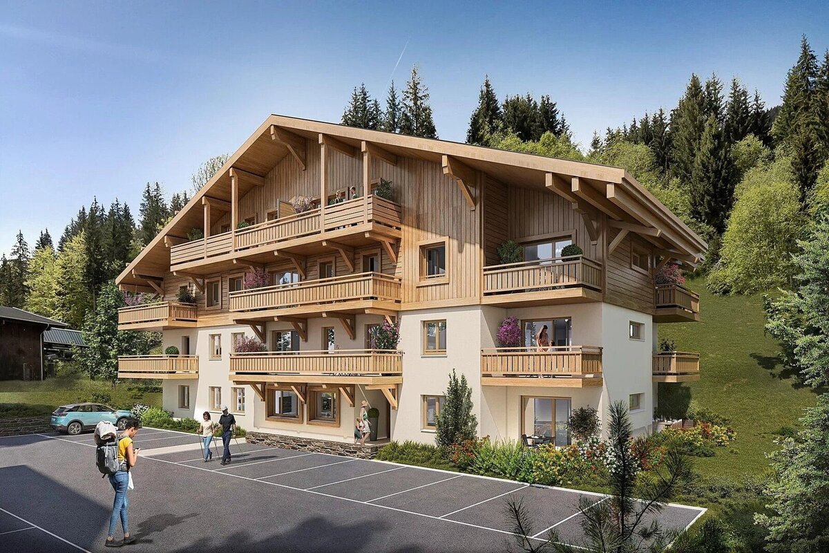 A large, new chalet-style building with many wooden balconies, nestled in a forested mountain landscape. People are in the foreground parking area.