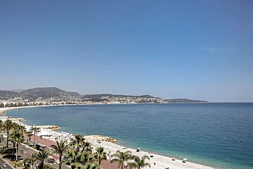 A sunny, high-angle view of a coastal beach with a palm-lined promenade, calm blue waters, and distant hills under a clear sky.