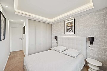 Modern bedroom with a white bed, upholstered headboard, grey patterned wallpaper, and light wood flooring. Features a built-in wardrobe and recessed lighting.