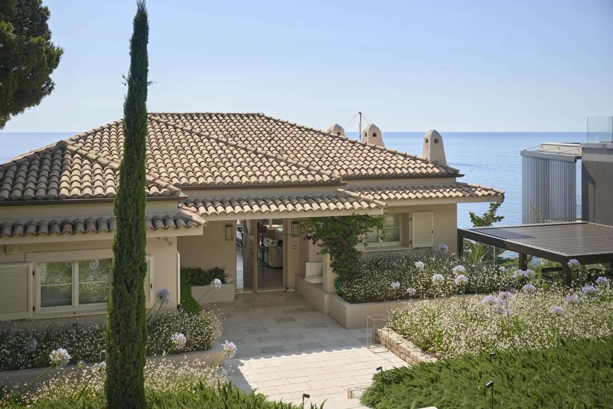 A Mediterranean-style house with a terracotta roof, surrounded by a vibrant garden and a tall cypress, with the calm blue sea in the background.