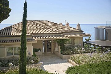 A beautiful Mediterranean-style house with a tiled roof, a tall cypress tree, and a lush garden overlooking the serene blue sea under a clear sky.