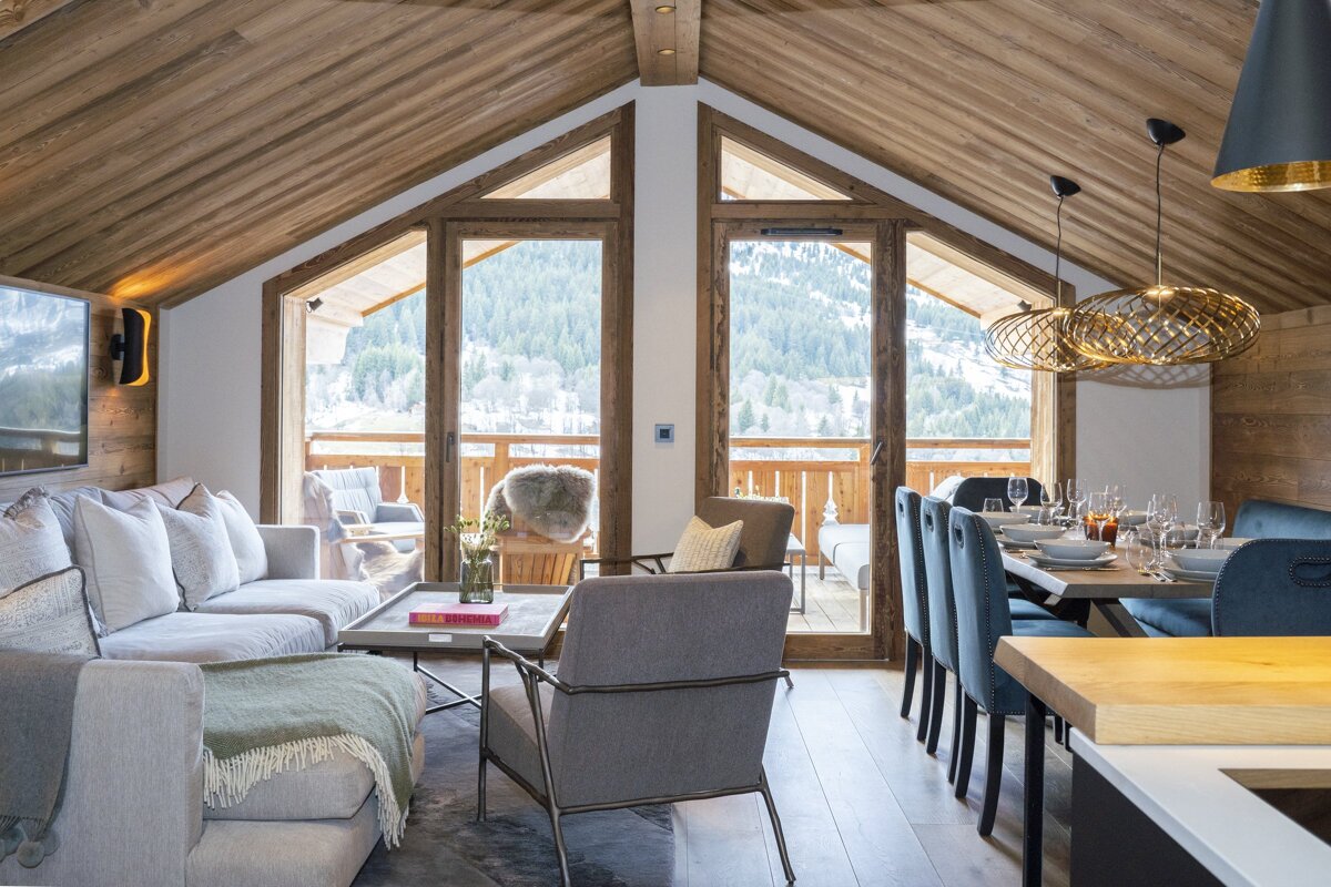 Luxurious open-plan chalet living and dining room with a pitched wooden ceiling, large windows, and a stunning view of snowy mountains.