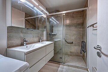Modern bathroom featuring a double vanity, illuminated mirrors, and a glass-enclosed walk-in shower with beige tiles.