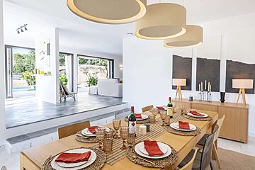 A modern, open-plan dining room with a fully set table, stylish decor, and large windows revealing a lush garden and pool area.