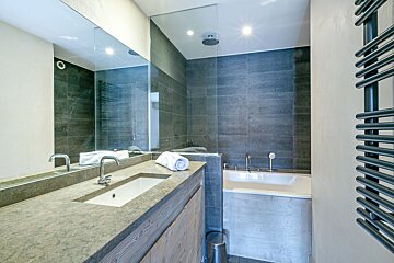 A modern bathroom with a long mirror, stone countertop, white sink, dark tiled shower/bath combo, and a heated towel rail.