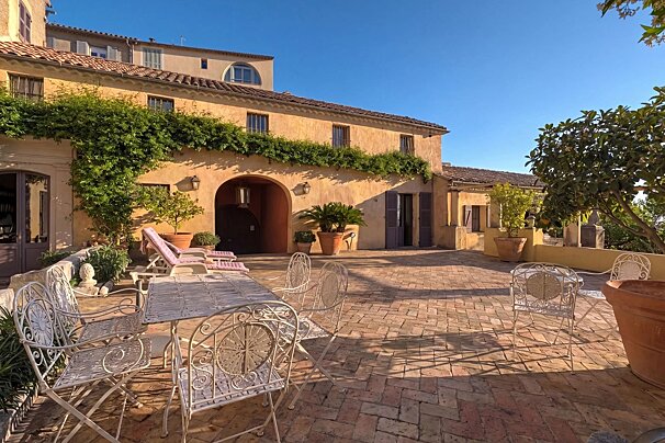 Sunny terracotta patio outside a charming Mediterranean-style villa with climbing vines, outdoor dining set, and lounge chairs under a clear blue sky.