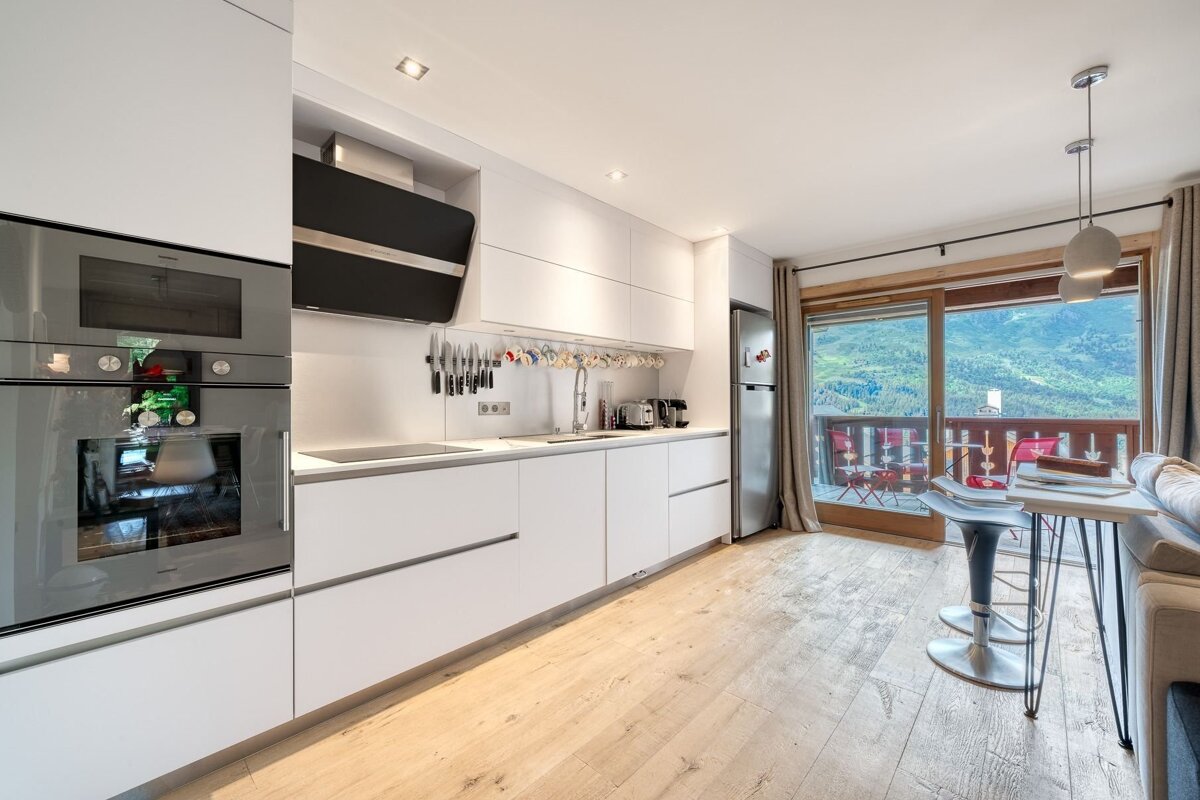 Modern white kitchen and living space with a large balcony offering panoramic views of green mountains and a valley.