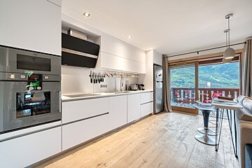 Modern white kitchen and living space with a large balcony offering panoramic views of green mountains and a valley.