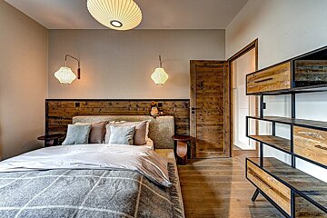 A rustic modern bedroom features a large bed, wooden headboard & door, unique pleated light fixtures, and an industrial-style metal & wood shelf.