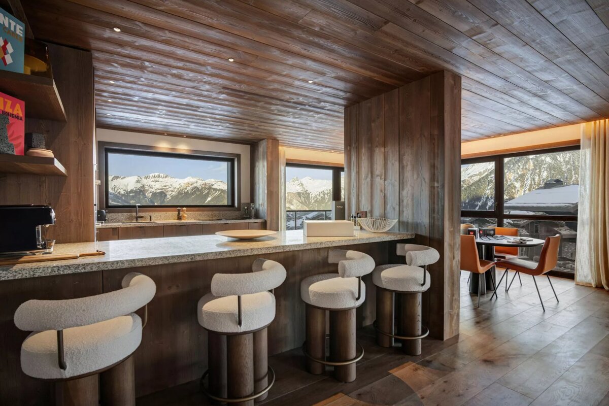 Modern, wooden chalet kitchen and dining area with stylish bar stools and expansive windows showcasing stunning snow-capped mountain views.