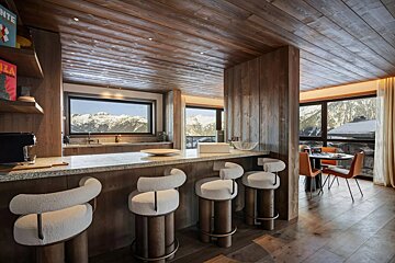 Modern, wooden chalet kitchen and dining area with stylish bar stools and expansive windows showcasing stunning snow-capped mountain views.