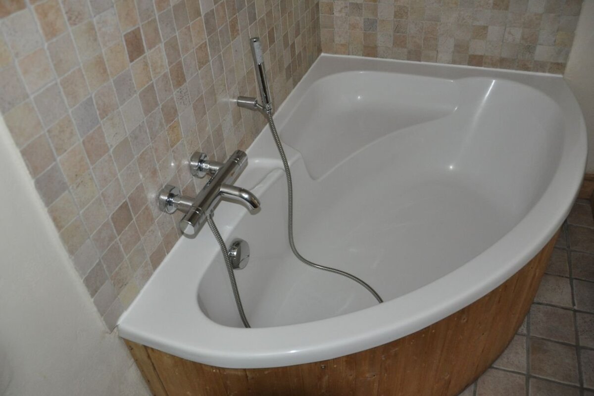 A white corner bathtub with wood paneling, chrome fixtures, and a hand shower, set against beige mosaic tiled walls.