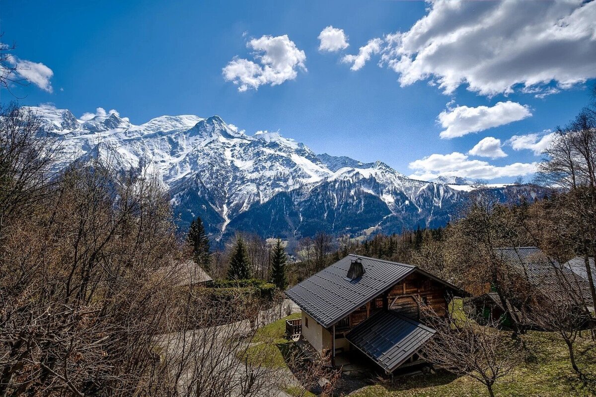 A cozy wooden chalet sits amongst trees, overlooking majestic snow-capped mountains under a vibrant blue sky with fluffy white clouds.