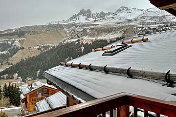 A snow-dusted chalet roof with a wooden railing overlooks a valley of evergreen forests and majestic snowy mountains under a cloudy sky.