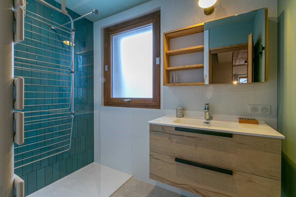 A modern bathroom features a teal-tiled shower with a drying rack, a wooden vanity with a sink, a mirror cabinet, and a window.