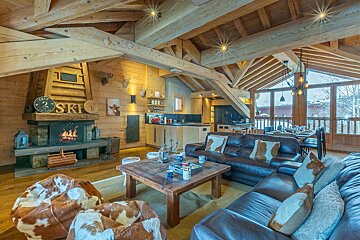 Cozy wooden chalet interior with a stone fireplace, leather sofas with cowhide pillows, bean bags, and exposed timber ceilings.
