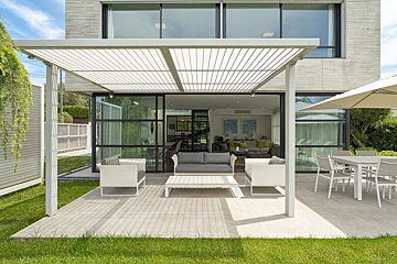 A modern house with a shaded outdoor patio featuring contemporary lounge seating, a dining area, and green grass. The interior living space is visible.