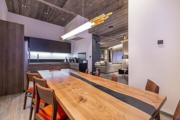 Modern open-plan kitchen, dining area with a long wood and resin table, and living room beyond. Features a textured wood ceiling and contemporary lighting.