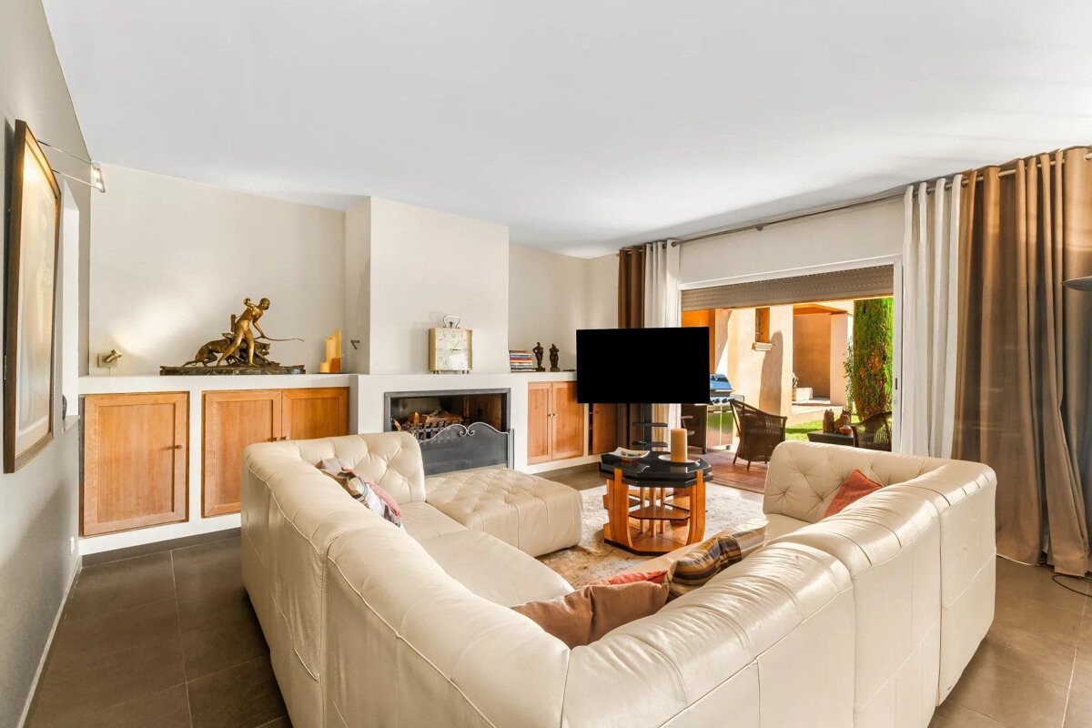 A bright living room features a large white sectional sofa, a fireplace, a TV, and opens to a sunlit outdoor patio.