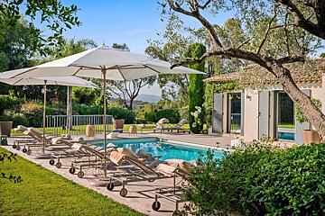A sunny swimming pool area with lounge chairs, white umbrellas, and a nearby stone house surrounded by lush greenery.